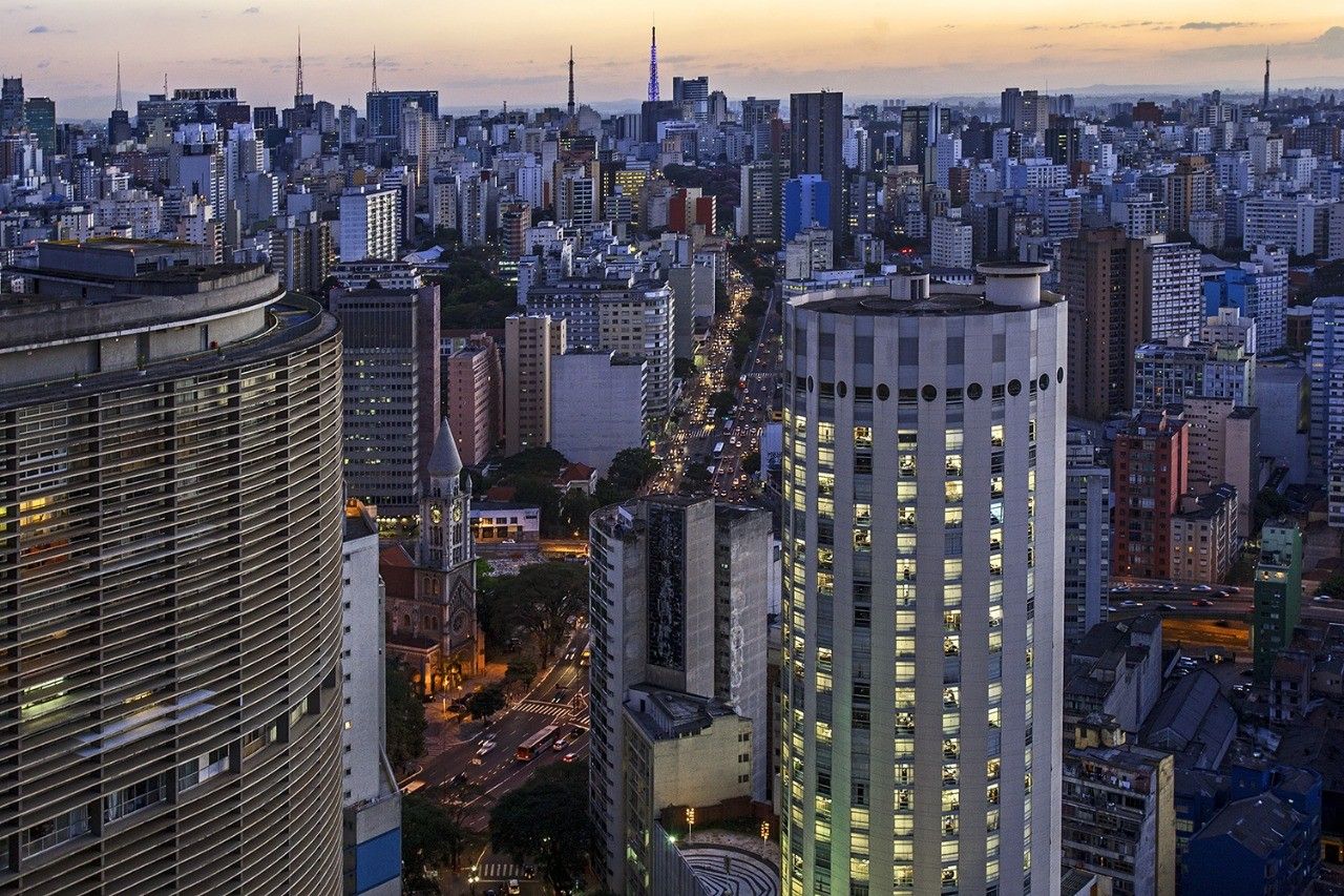 Vista aérea de São Paulo ao entardecer, com prédios iluminados e trânsito na avenida.