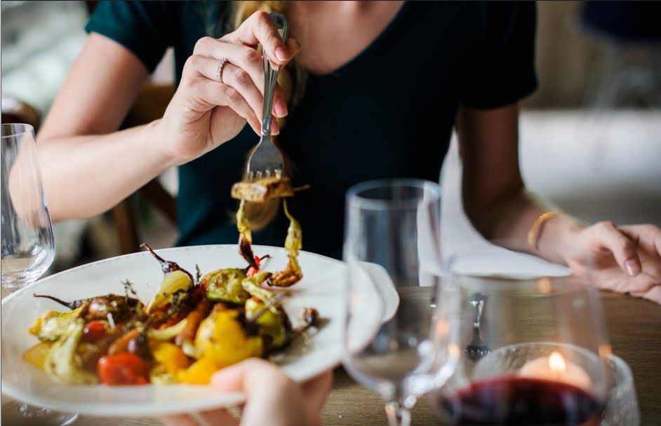 Pessoa jantando legumes assados em ambiente com luz baixa e vinho na mesa.