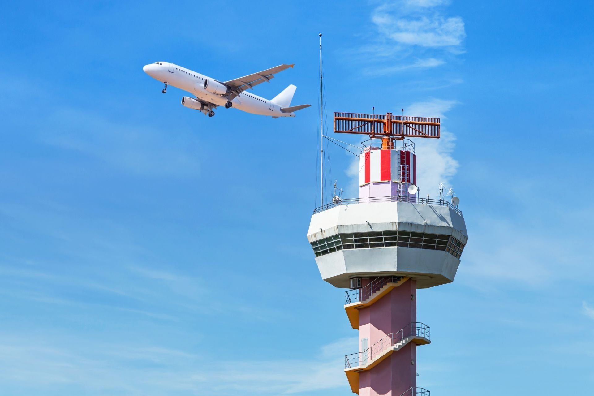 Torre de controle de tráfego aéreo sob um céu azul, com avião comercial ao fundo. Reprodução: Gizmodo