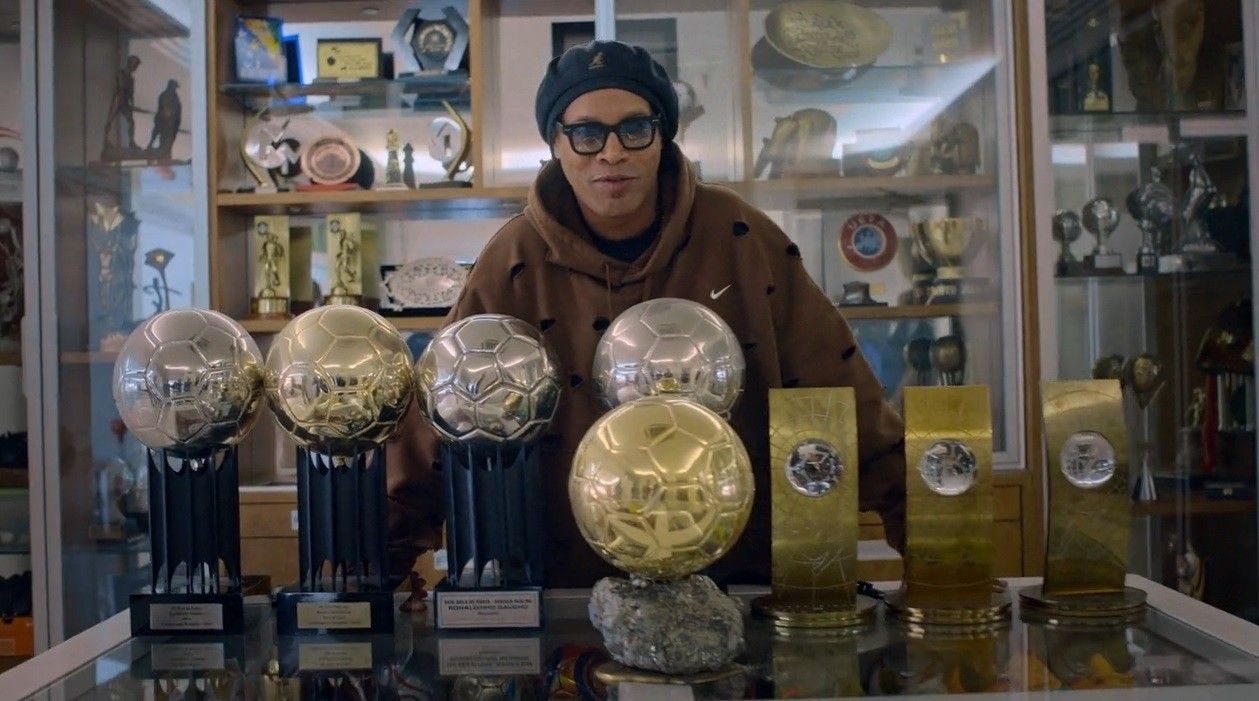 Ronaldinho Gaúcho posa com seus troféus em ambiente repleto de premiações.