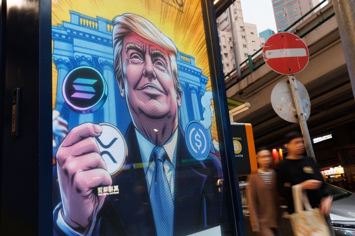 Pessoas passam por um anúncio de Donald Trump com referências a criptomoedas como Bitcoin e XRP. Legenda da imagem. Reprodução: Gizmodo