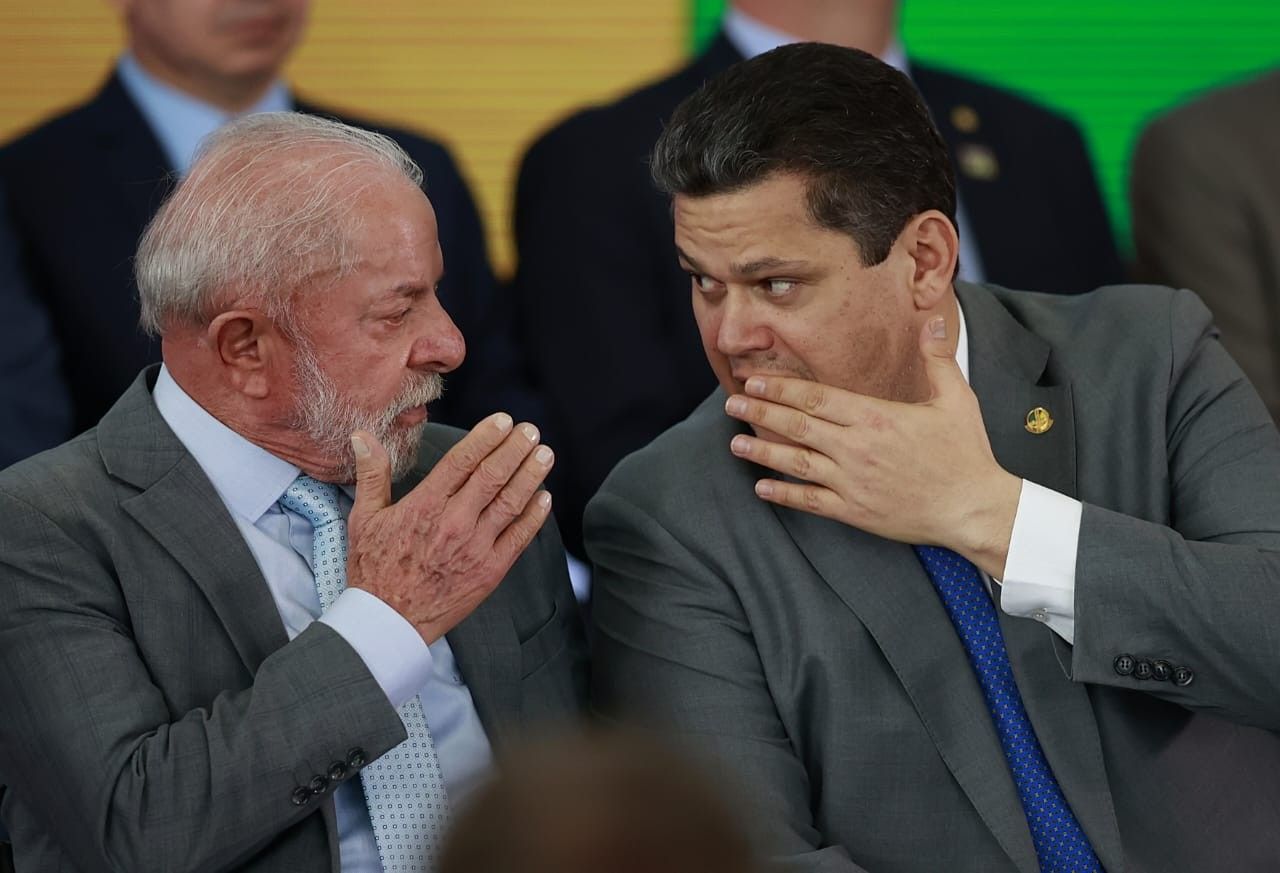 Dois homens conversam reservadamente durante evento formal, ambos com mãos próximas à boca.