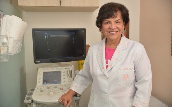 Médica sorri ao lado de aparelho de ultrassom em consultório clínico.