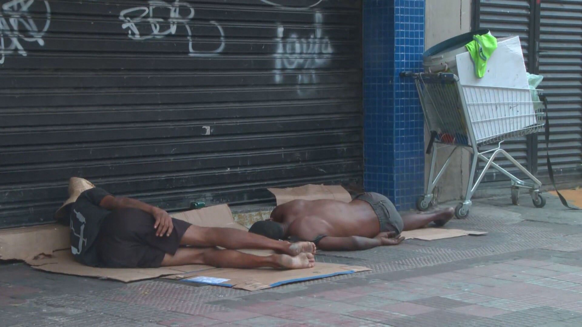Duas pessoas em situação de rua dormem sobre papelão em calçada urbana.